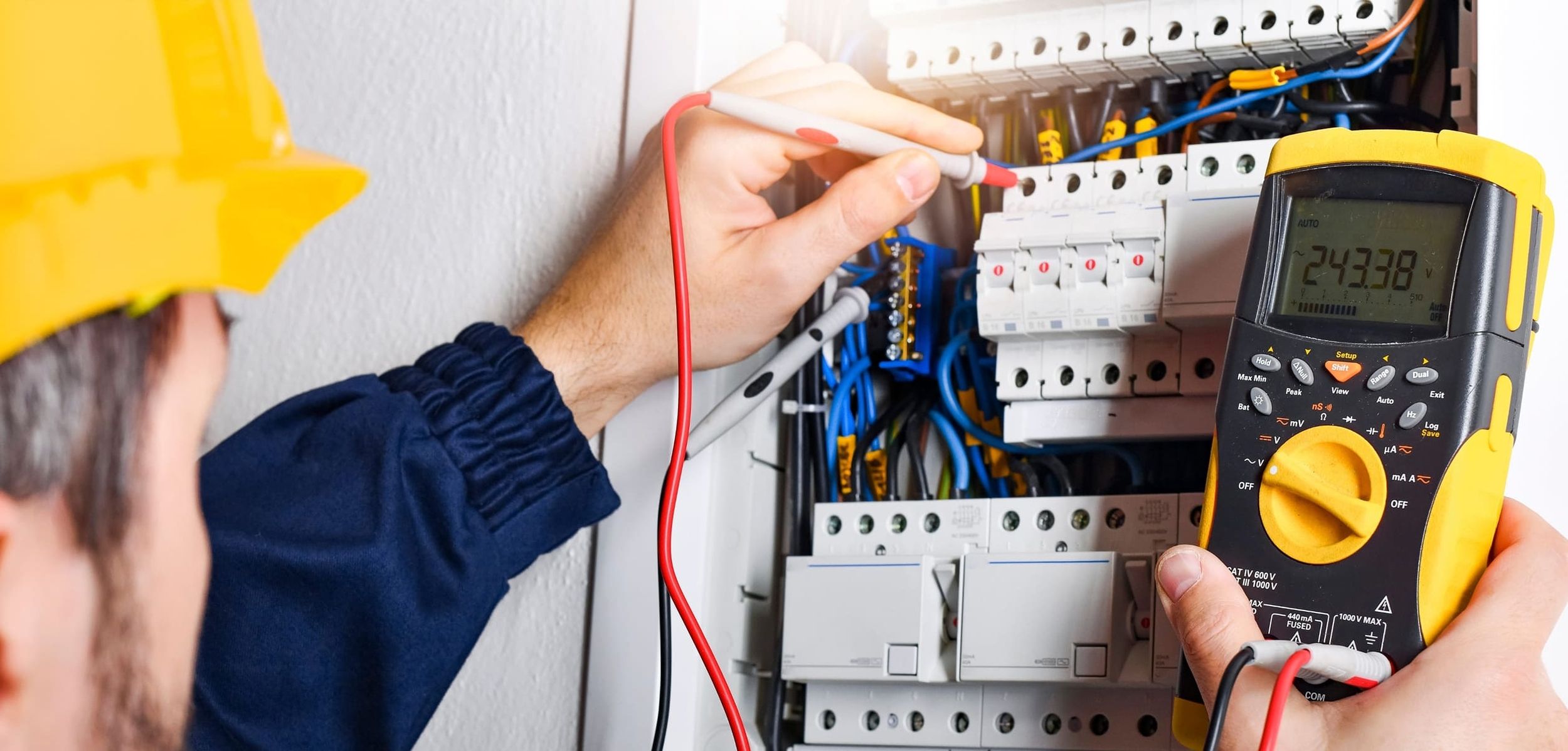 Elektriker in gelbem Schutzhelm prüft Sicherungskasten mit digitalem Multimeter